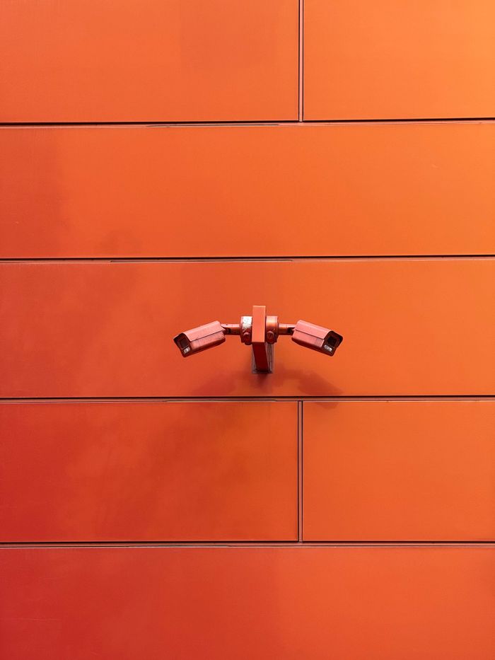 minimalist-orange-wall-with-cctv-cameras-33988300 Two CCTV cameras mounted on a vibrant orange wall, showcasing modern surveillance.